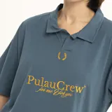 Remera tipo polo de manga corta, color verde oscuro, con cuello y abertura frontal. Presenta un estampado dorado en el pecho con el texto "PulauCrew" y un pequeño logo de corona de laurel bordado.