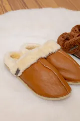 Pantufla de cuero color habano, forrada internamente con piel sintética (borrego) en tono crema. Posee costuras reforzadas y suela plana de goma.