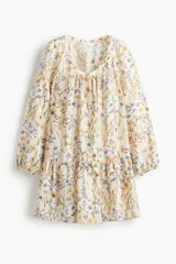 Vestido túnica evasé corto de color beige con estampado floreado en tonos azules y rosados. Cuenta con escote redondo amplio, botones forrados al frente y mangas raglán globo largas con puños elásticos. Presenta un diseño holgado con fruncido escalonado bajo la cadera.