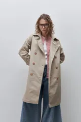 Trench clásico color marrón, de silueta relajada, confeccionado en tejido liviano, con cuello de solapa, doble abotonadura frontal y mangas largas con presillas regulables en los puños.