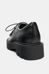 Zapato tipo oxford de cuero negro con cierre de cordones, costuras decorativas en la puntera y suela gruesa dentada de estilo track.