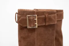 Bota de caña alta confeccionada en cuero gamuza color marrón, con diseño de punta fina y detalle de hebilla metálica en la parte superior de la caña.