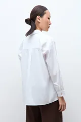 Camisa blanca de manga larga con cuello clásico y botones frontales. Presenta dos bolsillos asimétricos en el pecho, uno con solapa y otro sin.