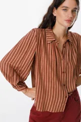 Camisa de manga larga con diseño de rayas verticales en tonos tierra, bordó y crema. Presenta cuello clásico, cierre frontal con botones y mangas con volumen y puños ajustados.