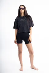 Remera de corte boxy fit, color negro con efecto lavado, de manga corta y cuello redondo. Presenta un logo bordado en el centro del pecho dentro de un óvalo.