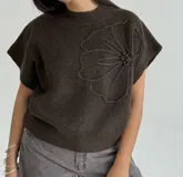 Chaleco tejido de punto color crudo, con cuello redondo y diseño de flor en relieve en el frente. Presenta un corte holgado y mangas cortas con terminación acanalada.