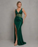 Vestido largo verde oscuro de satén, con escote en V y diseño de corset transparente en el torso con detalles de pedrería y varillas visibles. Cuenta con una falda larga de corte sirena con fruncidos en la cadera y una profunda abertura lateral en la pierna.