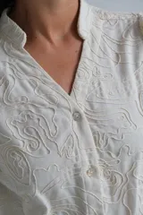 Blusa de color beige con cuello mao y cierre frontal de botones. Presenta mangas largas abullonadas con puños ajustados y un diseño con bordados en relieve de patrones orgánicos en toda la prenda.