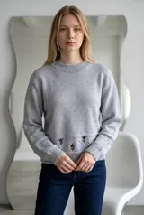 Sweater de punto color crema con cuello redondo, manga larga y diseño de calados en los puños. Presenta un detalle de bordado floral en la parte inferior y una franja con perforaciones decorativas.