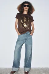 Camiseta de manga corta y cuello redondo, color marrón oscuro, con estampado frontal metalizado en tonos dorados que simula figuras de cerámica o arte tribal, y texto inferior que dice "Echoes in Gold".