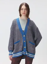 Cardigan de tejido de punto grueso en color gris con textura acanalada. Presenta un diseño oversize con cuello en V, cierre frontal de botones y detalles en color azul vibrante en el cuello, los bolsillos y los puños. Los puños y el ruedo incluyen franjas horizontales en blanco y azul.
