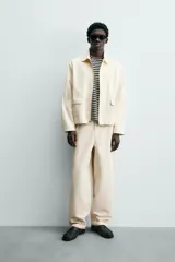 Pantalón chino de algodón color beige, de corte relajado (relaxed fit) y tiro medio. Presenta bolsillos delanteros y bolsillos traseros de vivo con cierre de cremallera y botón.