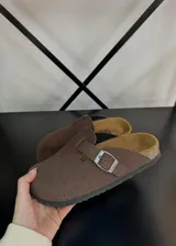 Zuecos de gamuza color marrón chocolate, con diseño de punta cerrada, plantilla anatómica y tira ajustable con hebilla metálica lateral.