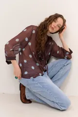 Blusa marrón semitransparente con estampado de lunares celestes, mangas largas abuchonadas, cuello en V con lazos y detalles de volados frontales.