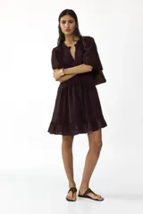 Vestido corto de color bordó, con escote en V y cuello de volados, mangas cortas abullonadas y botonadura frontal con alforzas e inserciones de encaje. Presenta costuras angulares con frunces en la cadera y falda con ribete de volado en el bajo.