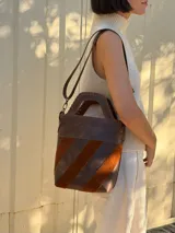 Bolso tipo matera confeccionado en cuero vacuno, con diseño estructurado y manija superior integrada. Presenta un cuerpo con franjas diagonales en gamuza de colores contrastantes (amarillo, verde y azul) sobre una base de cuero marrón, con detalles de costuras artesanales en los bordes.