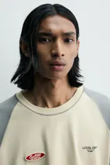 Camiseta de corte relajado con mangas ranglán en color gris y cuerpo en color beige. Presenta cuello redondo, costuras a contraste y detalles de parches y bordados en el pecho.