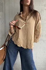 Camisa de lino color camel con cuello clásico, cierre frontal de botones y mangas largas. Presenta un delicado bordado floral tono sobre tono en las mangas y en el ruedo, el cual tiene un acabado festoneado.