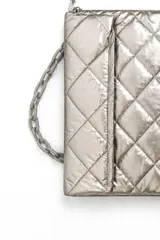 Cartera acolchada tipo bandolera de color plateado, con diseño matelasseado en rombos, cierre superior y una cadena de eslabones plásticos decorativa.