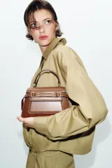 Bolso de mano tipo city color marrón, con diseño estructurado, costuras a la vista en color blanco y detalles de hebillas metálicas decorativas en la parte frontal. Cuenta con asa de mano superior, correa ajustable para usar como bandolera y cierre mediante solapa con imán.