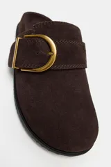 Zueco plano de serraje color marrón, con punta redonda y detalle de tira con hebilla metálica dorada en el empeine. Suela plana de goma.