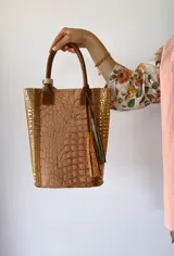 Bolso matero de cuero color beige con textura tipo piel de cocodrilo, detalles en cuero color marrón y borlas decorativas.