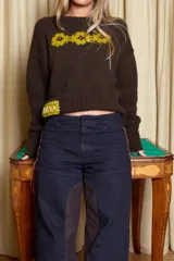 Sweater de lana merino color chocolate, tejido a dos agujas con diseño de flores bordadas en color amarillo en el pecho y parche decorativo con texto en la parte inferior.