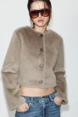 Chaqueta corta de cuello redondo y manga larga, confeccionada en tejido sintético con acabado efecto pelo. Presenta cierre frontal con botones a tono.