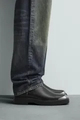 Bota tipo Chelsea de cuero negro con paneles elásticos laterales, suela de goma con volumen y detalle de pieza metálica en la puntera.