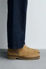 Bota estilo Chelsea de hombre, color marrón, fabricada en piel con acabado serraje (gamuza). Presenta elásticos laterales oscuros y suela de goma a tono con tacón bajo.