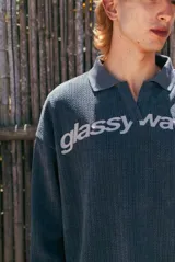 Buzo gris de tejido de punto con textura acanalada, cuello polo y escote en V. Presenta un estampado frontal con la inscripción 'glassy wa'.