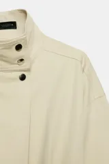 Mono largo de popelín color beige, con cuello subido, manga larga, bolsillos laterales ocultos y cierre frontal con cremallera y botones a presión.