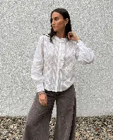 Camisa blanca de estilo romántico con detalles de alforzas, aplicaciones de puntilla y volado lateral en el frente. Presenta cuello con terminación de volado, mangas largas con puños abotonados y cierre frontal de botones.