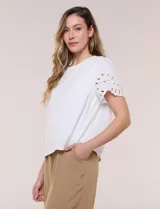 Blusa blanca de corte recto con cuello redondo y mangas cortas que presentan detalles de bordado calado y terminación festoneada.