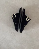 Zueco tipo sandalia de estilo jaula, confeccionado en gamuza negra con tiras entrelazadas y hebilla metálica dorada lateral. Cuenta con plantilla acolchada con logo de la marca.