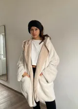 Abrigo tipo cárdigan de peluche en color crudo, con diseño holgado, capucha amplia y mangas largas con puños versátiles. Presenta una textura suave y mullida, ideal para un estilo casual y confortable.