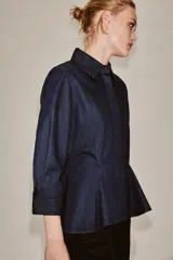 Camisa de denim azul oscuro con cuello solapa, manga larga con puños y diseño peplum en el bajo. Presenta cierre frontal con botones ocultos por solapa.