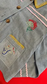 Chaqueta de jean celeste con bordados artesanales y a máquina. Presenta detalles de pespuntes ondulados en rosa y amarillo en el cuello, puños y bolsillos. Los bolsillos frontales tienen bordados de un limón y una figura marina estilizada.