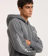 Saco con capucha y cierre frontal de color gris grafito, confeccionado en mezcla de algodón y poliéster. Presenta cordón ajustable en la capucha, puños y cintura ajustados, y una pequeña estampa localizada en el lado izquierdo del pecho con texto y tres círculos.