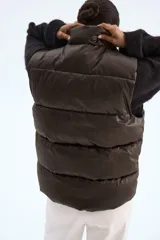 Chaleco acolchado tipo puffer color marrón oscuro, con cuello elevado, cierre frontal oculto por solapa con botones a presión y bolsillos tipo parche en el frente.