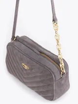 Bandolera de gamuza gris con diseño matelaseado en forma de V. Presenta un aplique metálico dorado en forma de labios en el frente y una correa combinada con cadena metálica dorada.