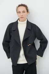 Chaqueta de paño color gris oscuro, con solapas amplias y cierre frontal mediante un alamar único. Presenta bolsillos laterales y mangas largas con puños rectos.