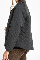 Campera negra de diseño capitoneado con cuello de solapa en contraste color marrón. Presenta cierre frontal con cremallera y botones a presión, mangas largas con puños ajustables y un corte recto.