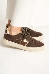Zapatillas bajas de gamuza color marrón con cordones y suela de goma color crema. Presentan una franja lateral con estampado de vaca y el logo de la marca bordado en el lateral.