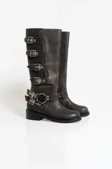 Botas de caña alta confeccionadas en cuero con efecto gastado. Presentan un diseño de estilo biker con múltiples hebillas laterales y apliques metálicos, incluyendo una argolla decorativa en el tobillo. Cuentan con un taco bajo cuadrado y suela de goma.