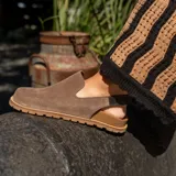 Zueco de cuero gamuzado color marrón, con diseño destalonado, tira elástica trasera y suela plana con plataforma dentada.