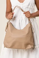 Cartera tipo hobo de ecocuero color beige, con textura granulada, cierre superior con cremallera, doble asa corta y correa larga regulable con cadena decorativa.