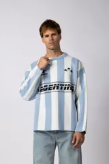 Sweater de tejido de punto con diseño a rayas verticales en color celeste y blanco. Presenta un corte holgado, cuello redondo y detalles gráficos en negro en el frente, incluyendo la palabra 'Argentina' y estrellas bordadas.