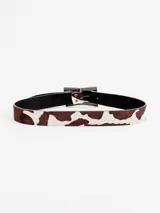 Cinto de cuero con estampado tipo animal print de vaca en tonos marrón y blanco, con hebilla metálica rectangular envejecida.