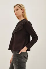 Camisa de manga larga color negro, con cuello redondo y cierre frontal de botones. Presenta un diseño con volados en la parte superior delantera y detalles de encaje bordado en los hombros y pecho.